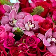 Elegant floral arrangement with red and pink roses, burgundy cymbidium orchids, and greens in a tall pedestal glass vase